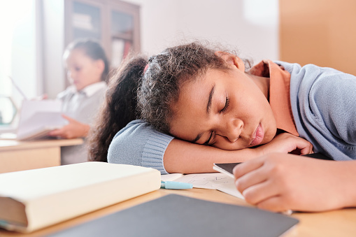 Exhausted or bored schoolgirl in casualwear keeping her head on desk while napping at lesson on background of classmate with book