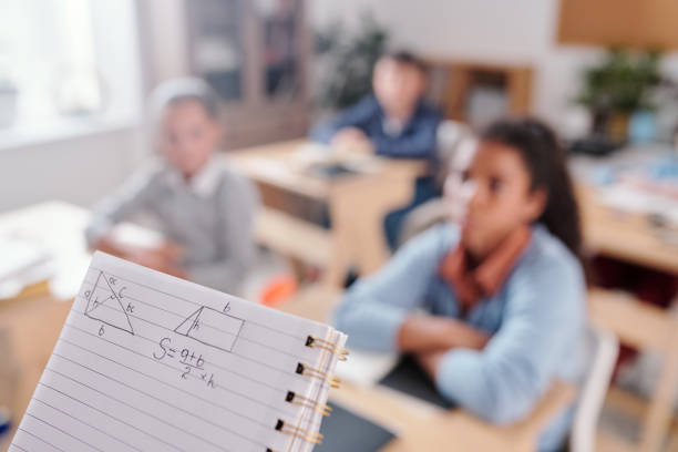 Copybook with geometric shapes and formula against pupils sitting by desks in rows and listening attentively to teacher at geometry lesson