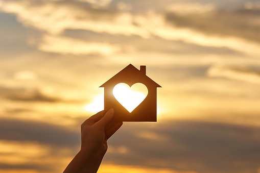 Woman hand holds wooden house in the form of heart against the sun