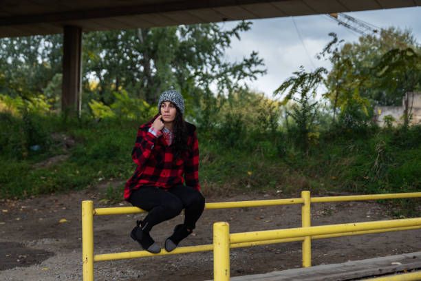 молодая депрессивная бездомная девушка или женщина, сидящая одна на заборе под мостом на улице в холодную погоду избирательного внимания - dirty grunge teenager relationship difficulties стоковые фото и изображения
