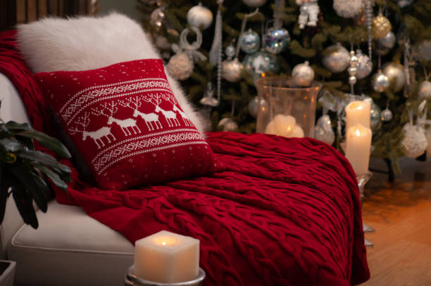 istock Christmas still life with gorgeous red knit blanket and deers pattern cushion on a couch, big candles on a floor and decorated Christmas tree on the back 1283218559