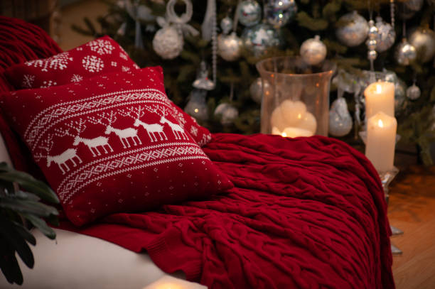 istock Christmas still life with gorgeous red cable knit blanket and deers pattern cushion on a couch, big candles on a floor and decorated Christmas tree on the back 1283218640