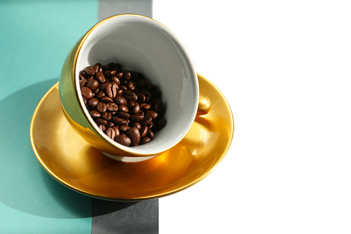 Front view for golden porcelain coffee cup copy with fresh roasted arabica beans. Large golden cup in antigravity position on the geometric grey and tiffany blue background.