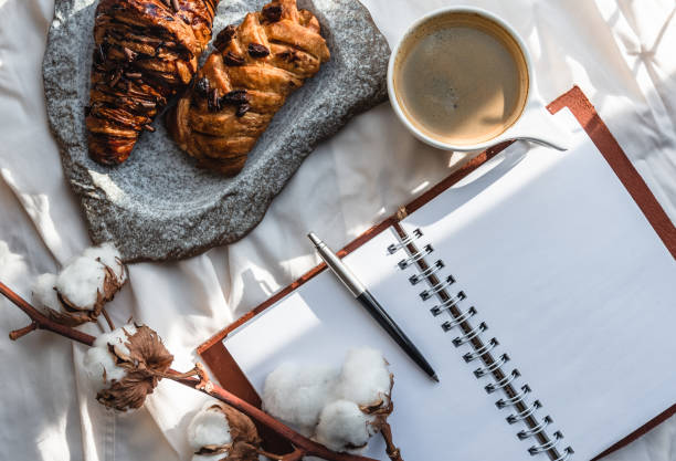 Coffee mug with croissant and empty notebook and pencil for business plan and design ideas onon white cloth from above, cozy and tasty breakfast, vintage toned