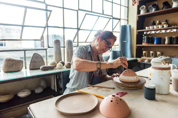 Portrait of senior female pottery artist painting a pottery bowl in her workshop