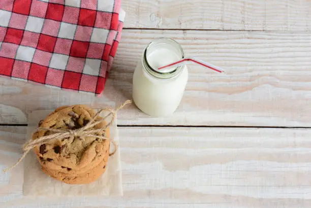 High angle shot of an after school snack of chocolate chip cookies and an old fashioned bottle of milk. The cookies are tied with twine and with a napkin on a rustic wood kitchen table.
