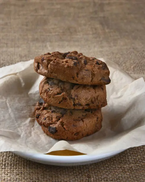 Closeup of three chocolate chocolate chip cookies on a plate. The delicious fresh baked treats and plate are on a burlap table cloth.