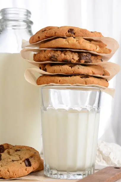 Closeup of a stack of fresh baked cookies on top of a glass of milk. Vertical format with a bottle of milk and flour in the background. Chocolate Chip, Oatmeal Raisin, White Chocolate cookies.
