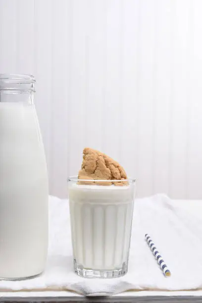 A glass of milk with a cookie dunked inside. A bottle of milk and drinking straw in a predominantly white kitchen.