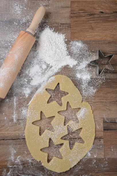 Top view of raw cookie dough with star shapes and cookie cutter on wood table with a rolling pin and flour sprinkles.