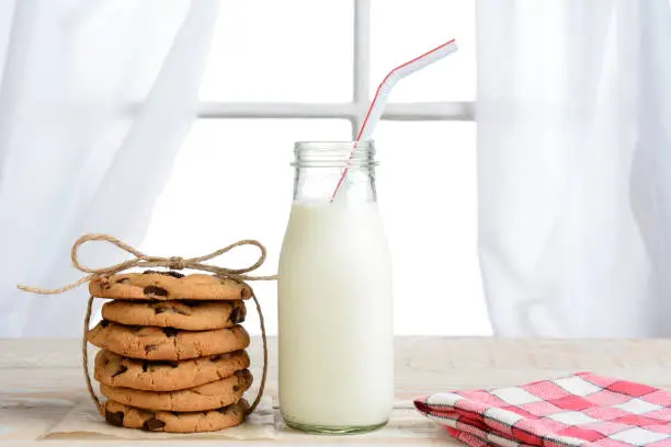 Horizontal shot of an after school snack of chocolate chip cookies and an old fashioned bottle of milk. The cookies are tied with twine and with a napkin on a rustic wood kitchen table.