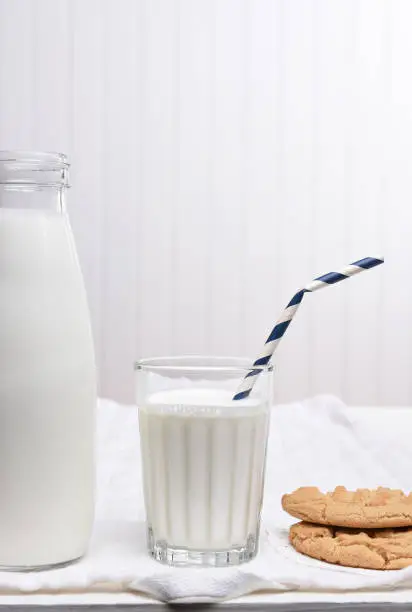 A Milk and Cookie Snack on a white table with white wall. The glass has a Drink Straw with cookies by its side.