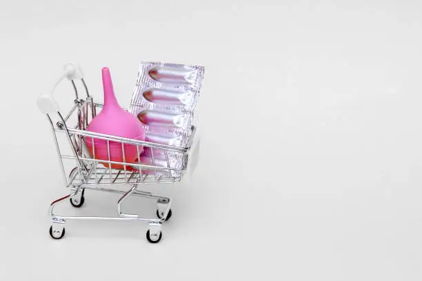 Medical suppositories in a silver blister pack and a pink rubber medical enema are placed in a toy shopping cart. Packaging of contraceptives, vaginal, rectal candles. A pink medical enema. Copy space.