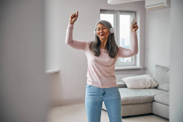 Charming woman listening to music and dancing in living room Beautiful lady in wireless headphones holding smartphone and smiling while moving to the rhythm of music at home 40 49 years stock pictures, royalty-free photos & images