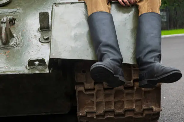 Fragment of the front of an old military tank. A track, an armor plate and the legs of a man in soldiers' boots are visible. Background.