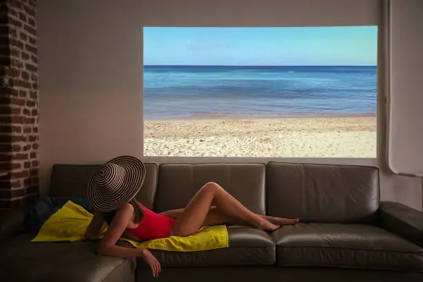 Woman pretends to sunbathe on a fake beach looking at the sea of a projector during a pandemic at home in an apartment