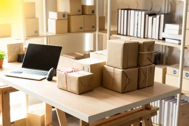 Photo of Parcel boxes on shelf and color shopping bags placing near laptop on table. SME business on shopping online at home office packaging on background is popular business.