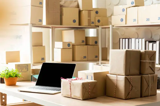 Photo of Parcel boxes on shelf and color shopping bags placing near laptop on table. SME business on shopping online at home office packaging on background is popular business.