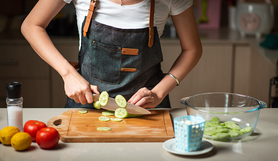 Woman slicing cucumber while making a vegetable salad at home. Healthy meal preparation in the kitchen