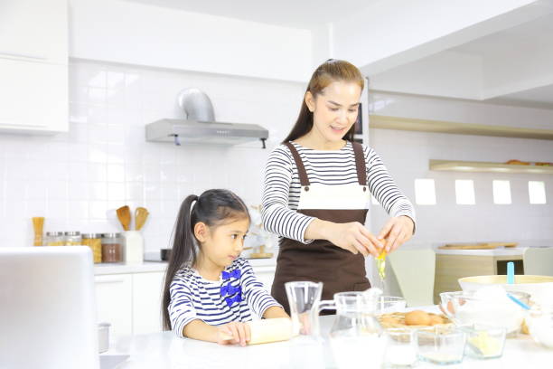 mãe asiática feliz ensinando sua filha jovem a assar pão na cozinha branca enquanto quebra ovo para misturar com farinha - bread food baked 7 grain bread - fotografias e filmes do acervo