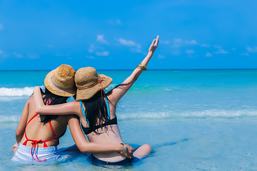 Happy young women travel on beautiful beach and look at beautiful sea, friend sitting together on tropical beach in long weekend holiday summer. Traveler girls in bikini and straw hats. enjoying life