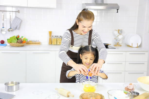 happy asian mother teaching her young daughter breaking egg for mixing with flour to bread baking in white kitchen - bun bread 7 grain bread dough imagens e fotografias de stock