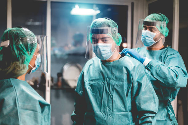 médicos y enfermeras preparándose para trabajar en el hospital para la operación quirúrgica durante el brote de pandemia de coronavirus - trabajadores médicos que se visten dentro de la clínica - enfoque en la cara del hombre derecho - traje protector fotografías e imágenes de stock