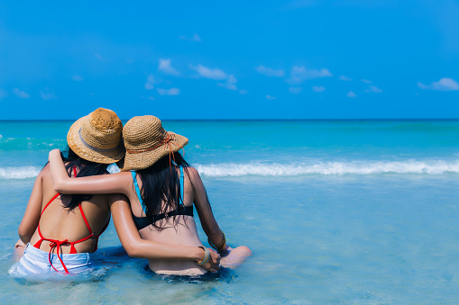 Beautiful young women enjoying on beach and look at nice sea, friend sitting and arms around neck, hug each other on tropical beach in summer vacation time. Traveler girls wear bikini and straw hats