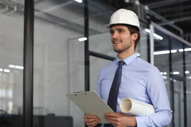 istock Confident young business man in hardhat holding blueprint and looking away while standing indoors. 1283314402
