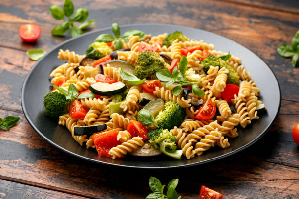 pâtes fusilli aux légumes, brocoli, courgettes, poivrons rouges, aubergines, tomates et basilic sur assiette noire - gourmet salad dinner prepared fish photos et images de collection