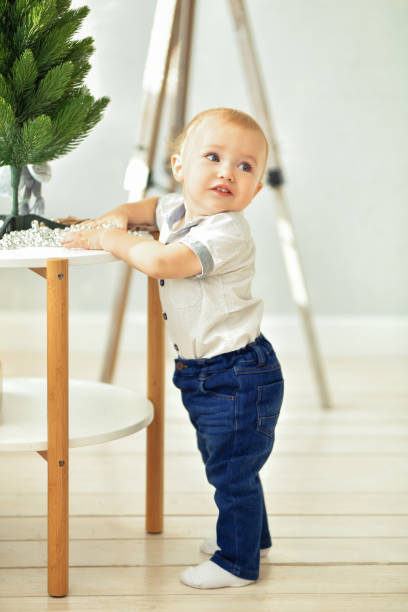 青いジーンズと白いシャツを着た小さな金髪の男の子がクリスマスツリー付きのテーブルに立っています。垂直撮影 - playground cute baby blue ストックフォトと画像