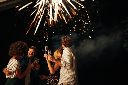 Four friends standing outside, toasting with glasses of wine and watching fireworks