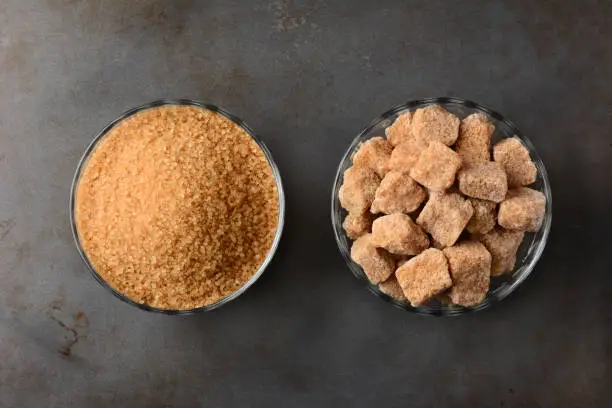Two bowls of brown sugar. One bowl of granulated raw turbinado sugar and a second bowl of natural brown sugar lumps. High angle view on a used metal baking sheet