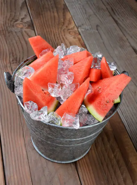 Closeup of a bucket filled with watermelon slices and crushed ice. The pail is sitting on a rustic wood picnic table. Vertical composition shot from a high angle.