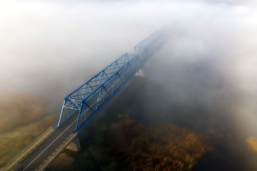 View from above on a metal bridge over the river Lielupe wrapped in fog in the autumn morning. The far end of the bridge disappears completely in fog and is not visible. Epic, fantastic, mystical view, soft focus