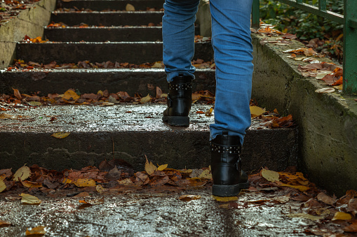 A girl in jeans and boots walks up the wet steps of a staircase covered with fallen autumn leaves in the rain. Selective focus. Copy space for your text. Rainy weather theme.