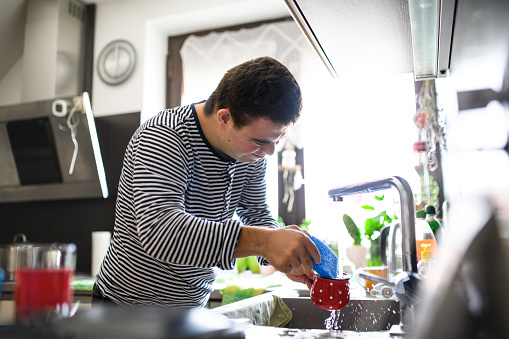 Portrait of down syndrome adult man standing indoors in kitchen at home, washing the dishes.