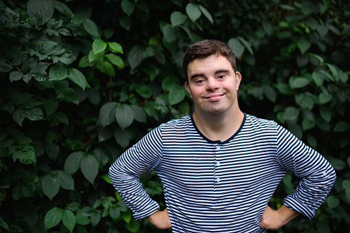 Portrait of down syndrome adult man standing outdoors at green leaves background, looking at camera.