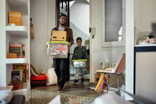 Young Boy Following Father into New Apartment on Moving Day Afro-Caribbean single father in late 20s and 7 year old son smiling as they carry cardboard boxes and plastic containers into new home. belongings photos stock pictures, royalty-free photos & images