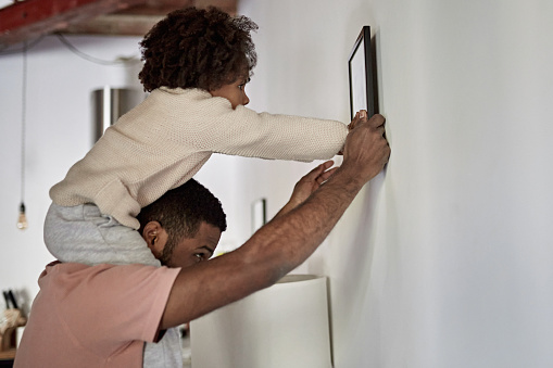 Side view of Afro-Caribbean man in late 20s with 3 year old daughter on his shoulders as they find a place to hang picture in new apartment.