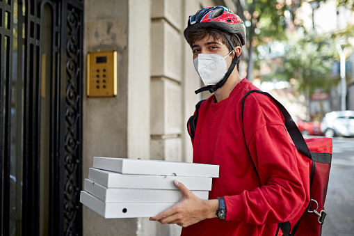 Candid portrait of bicycle messenger wearing KN95 face mask, helmet, insulated backpack, and looking at camera while holding pizza boxes for delivery.
