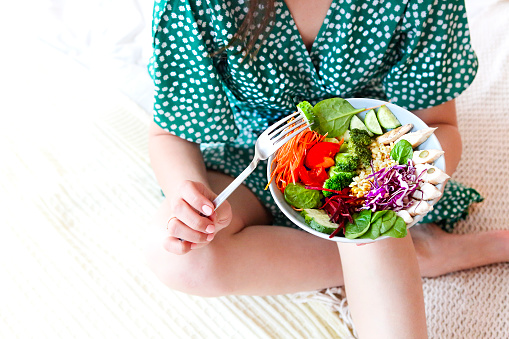 Top view of anonymous barefoot female in green dress showing bowl with healthy salad with vegetables and chicken meat. Close up