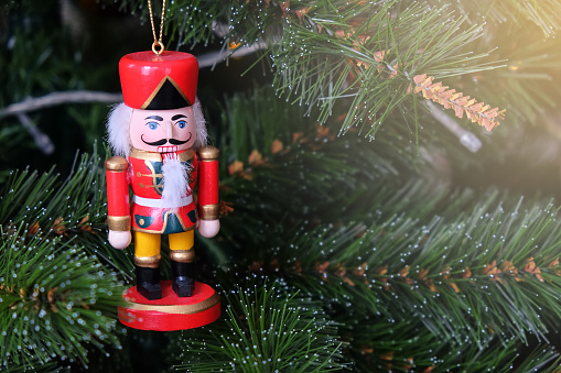 Toy funny nutcracker hanging on Christmas tree. New Year, holiday background with place for text and flare. Soft selective focus. Front view. Horizontal
