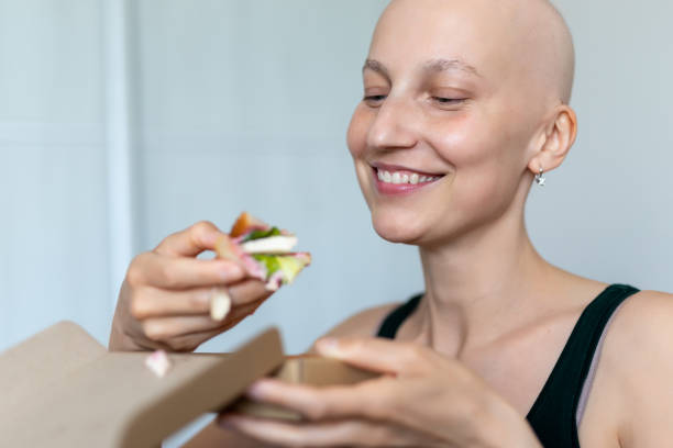 giovane adulto bellissimo caucasico calvo rasato capelli donna ritratto godere di mangiare panino per il pranzo a casa al coperto. persona sopravvissuta al cancro al seno nutrizione sana consapevolezza dietetica. alopecia chemioterapica - usare la bocca foto e immagini stock