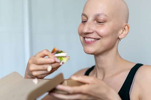 Young adult beautiful caucasian bald shaved hair woman portrait enjoy eating sandwich for lunch at home indoor. Breast cancer survivor person nutrition healthy diet awareness. Chemoterapy alopecia.