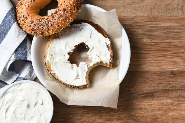 Bagel and Cream Cheese A bagel with cream cheese on a plate and a bite taken out. On a rustic wood table with copy space. cream cheese stock pictures, royalty-free photos & images