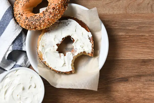 A bagel with cream cheese on a plate and a bite taken out. On a rustic wood table with copy space.