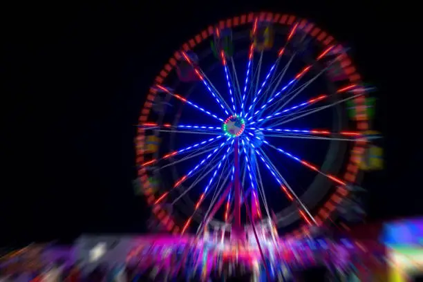 Radial blur of a ferris wheel at night. The ferris wheel is lit up red and blue. blurred color below. Black sky.