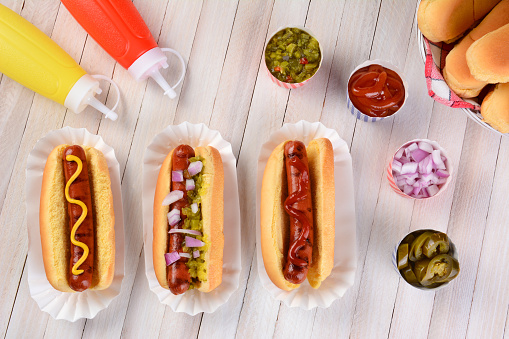 Overhead still life of a summer picnic table with hot dogs and condiments. Three franks in buns with ketchup, mustard, and relish surrounded by condiments.