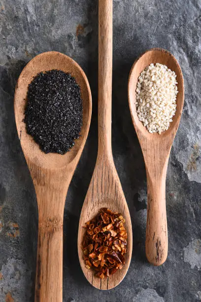 Top view of three wooden spoons with sesame seeds, poppy seeds, and dried chilies. Vertical format on a gray slate surface.